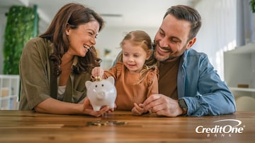 A young girl puts coins in her piggy bank with the help of her parents, who may be teaching her the importance of starting early to build wealth.